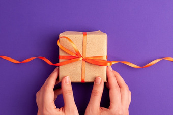 Zwei Hände halten Geschenk mit orangenem Band vor einem lila Hintergrund